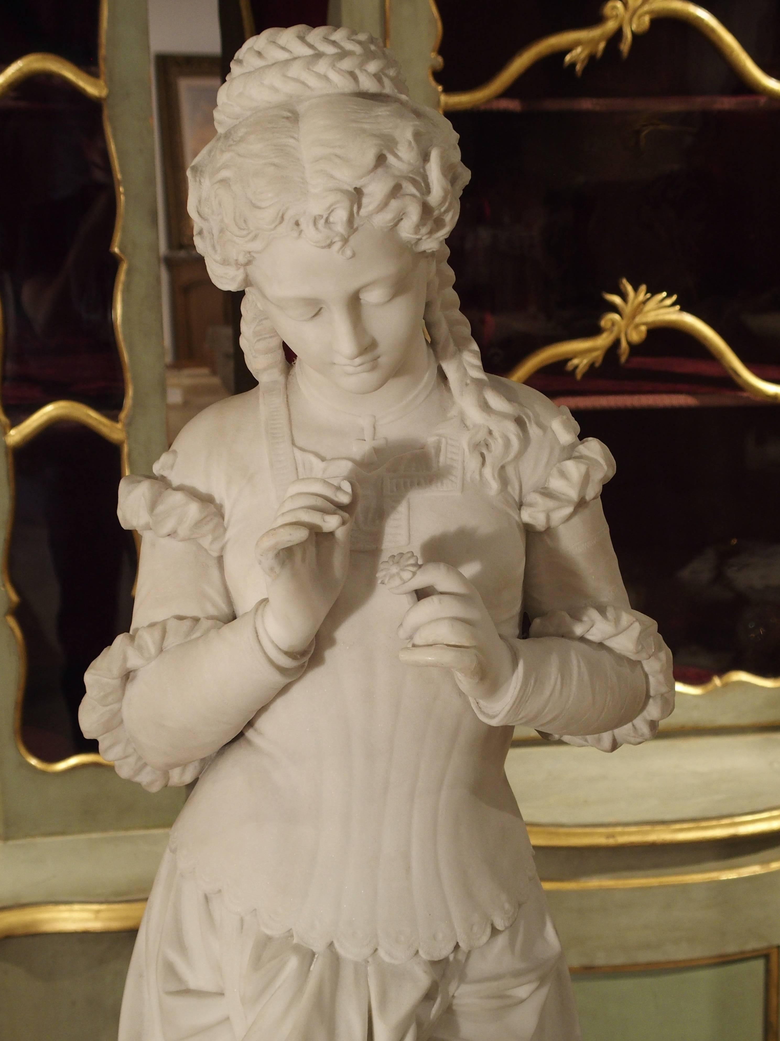 This beautiful marble statue of a young woman holding a single flower dates to the 19th century and is of the Italian school. She is shown wearing a long, dropped waist dress that is slightly hiked up by her purse. The back of the dress is cross