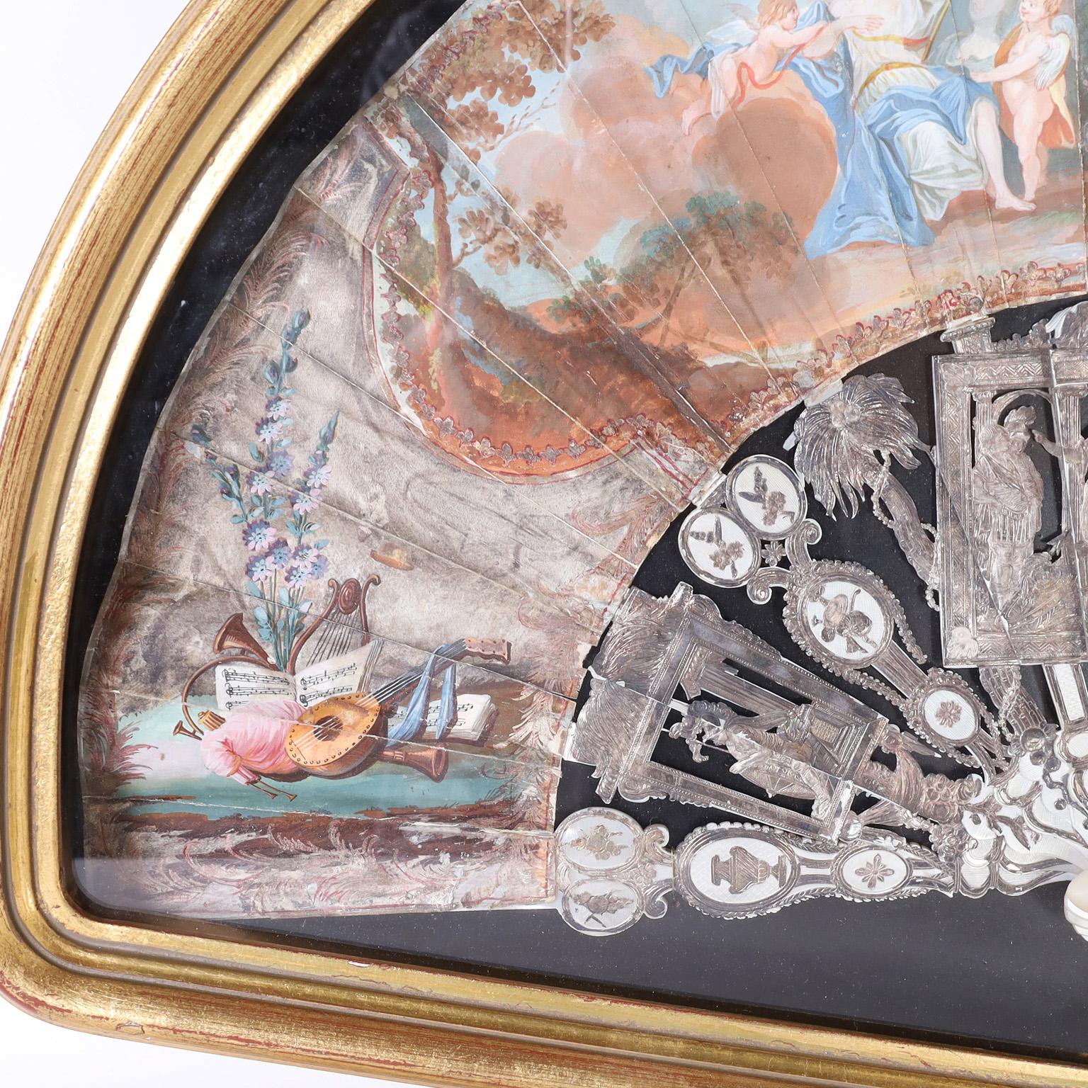 Impressionnant éventail à main italien du XIXe siècle, remarquablement conservé avec ses baguettes et ses protections en métal argenté représentant des dieux anciens, des palmiers et des emblèmes classiques. Les feuilles de papier sont des