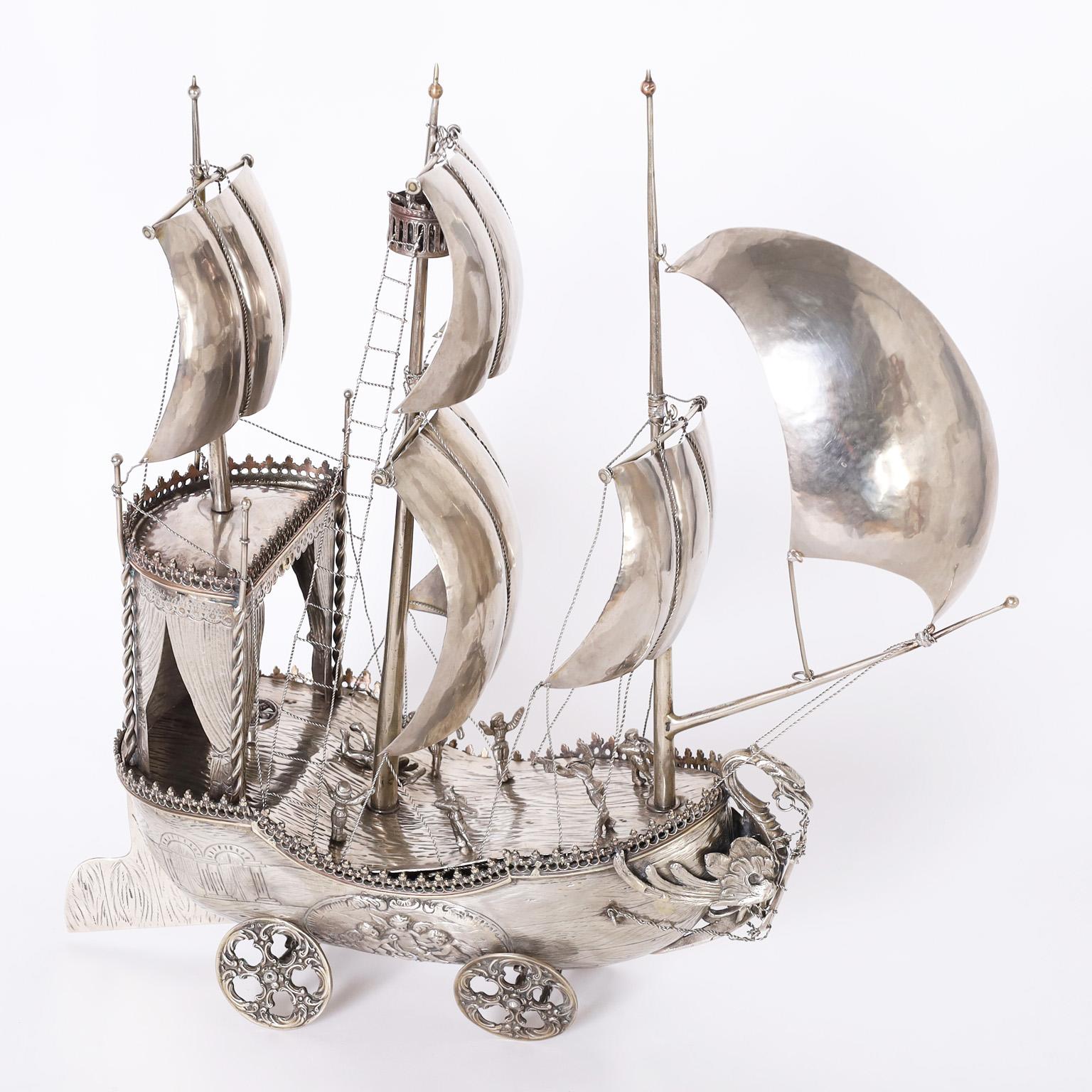 Enchanting antique sailing ship or nef hand crafted in silver plate on brass with plenty of unexpected details including hand hammered billowing sails, a crows nest, dramatic dragon figurehead, busy sailors embossed putis, and chariot style wheels.