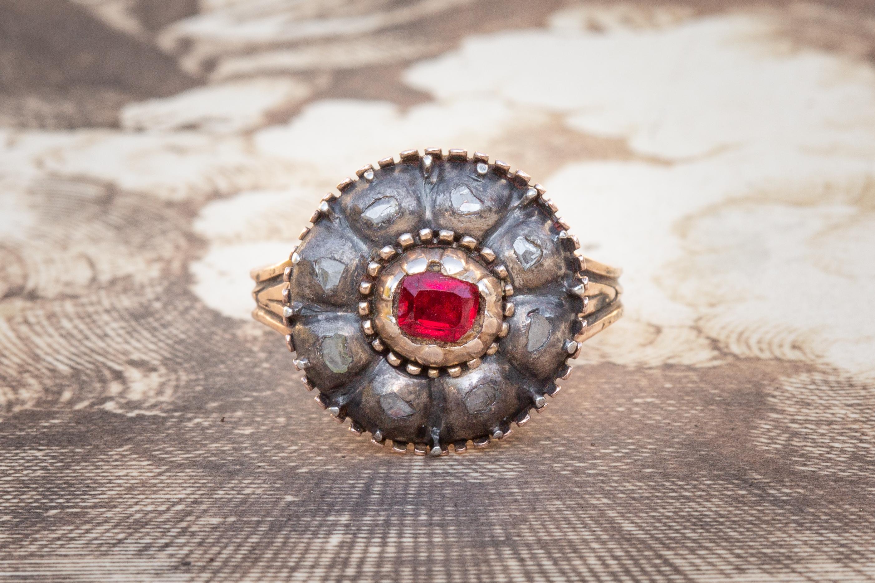 This lovely antique Italian cluster ring dates to around 1800 and is set with a garnet and diamonds. In the middle of the ring head lies a bright red garnet, surrounded by a cluster of 8 flat rose cut diamonds in rubover silver settings. The flower