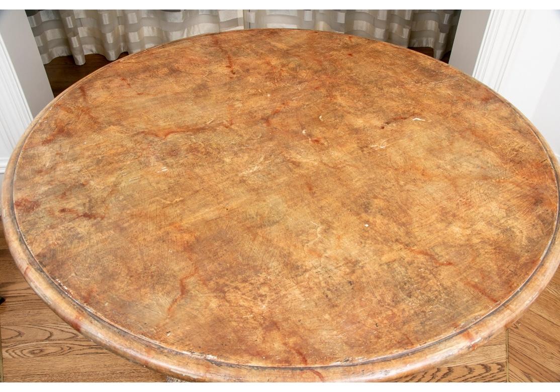 The large round tabletop painted in a mottled red-brown faux marble finish with a bull nose edge. Raised on a classical carved solid tripod base with faux marble painted panels and each decorated with a crest, a crown or a medallion. The carved