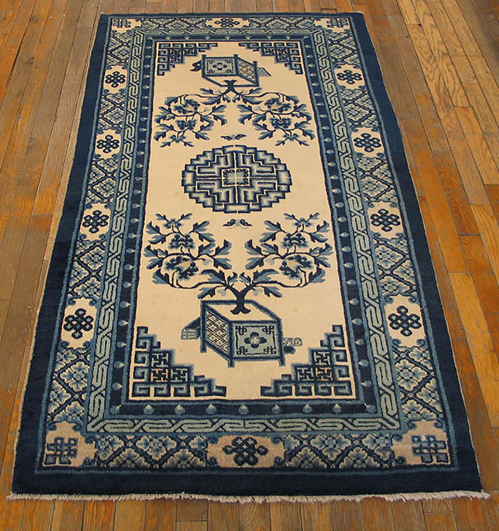 Blue and white palette with central fret medallion and split tree peony bushes growing from square vases above and below, framed by fret corners. The main border is in a knot and textile pattern with wave and pearl inner borders.
  