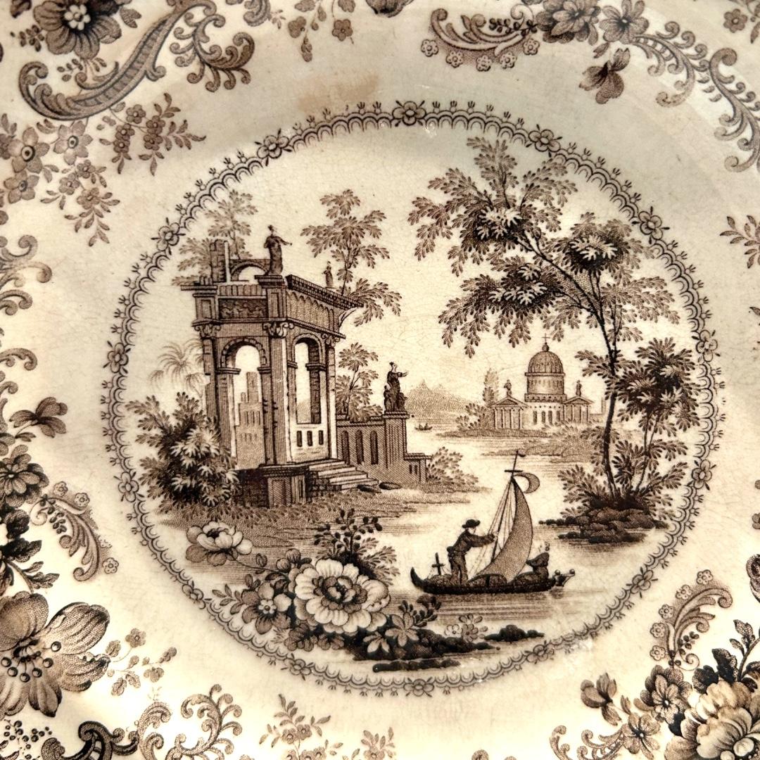 This elegant plate has a scalloped edge, floral design trim with old architectural buildings along a winding river with a sailboat in center; transfer printing, a decorative technique which was developed in England in the mid-18th century. J.Hall &