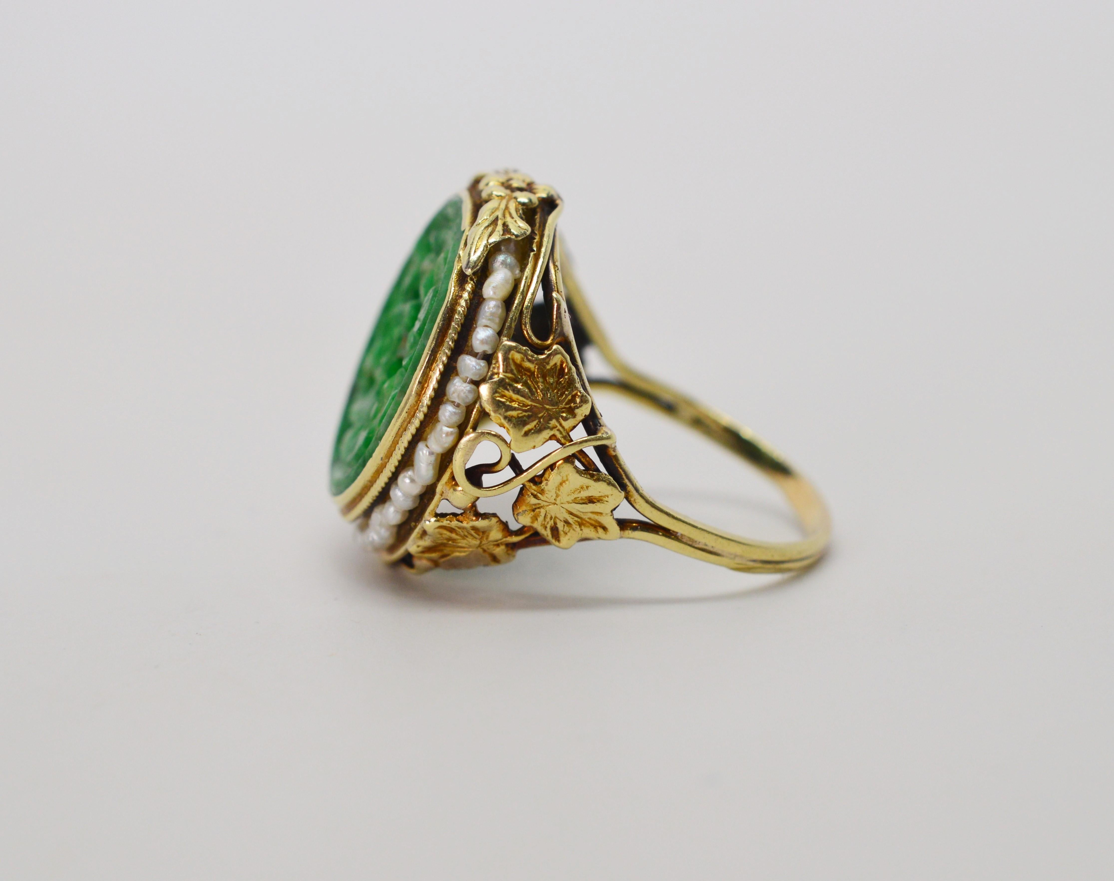 Artful leaves and vines in fourteen karat yellow gold intertwine on the shank of this hand-crafted ring which features a center of placket hand-carved sage green Jade accented with wreath of petite pearls. In ring size 7.5. 