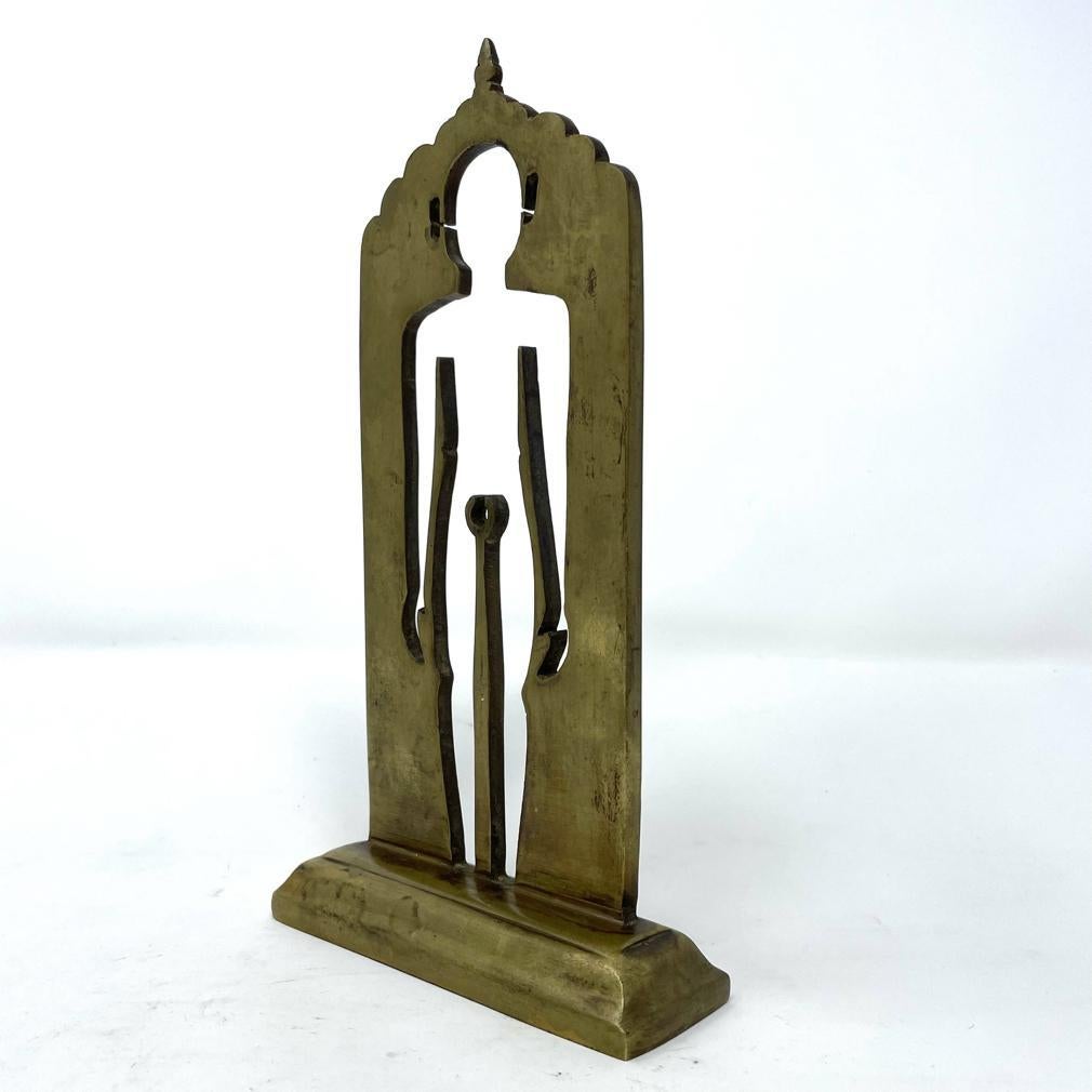 Antique Jain Siddhapratima Yantra. A striking brass votive image of the liberated soul, a male figure standing in meditation (kayotsarga) as a cutout of a thick piece of metal with finished base and scalloped roof line. The vacant image represents a