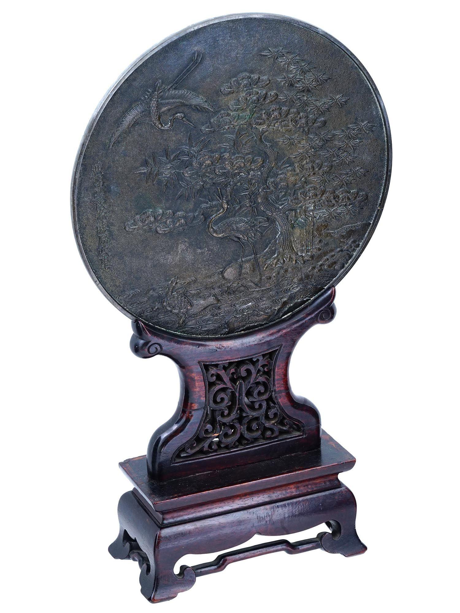 Antique (late 19th century) circular patinated bronze table screen depicting stylized standing crane in a lush setting with crane in flight above, mounted on carved openwork wooden stand, depicting.  Signed.  With silvered rear for use as a mirror.
