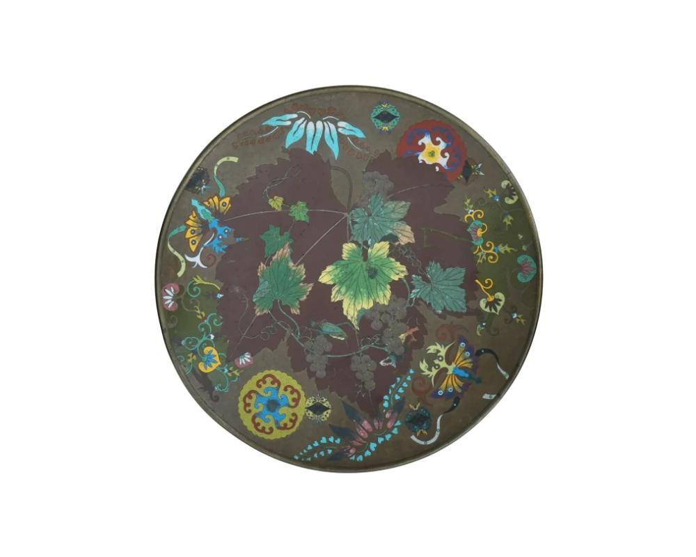 A large antique Japanese Meiji period, 1868 to 1912, cloisonne enamel metal charger plate depicting a multicolored picture of vines and grapes with a grasshopper and other plant motifs on a greenish brown ground.

The back side is adorned with a