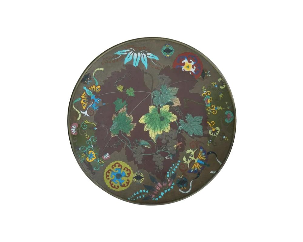 A large antique Japanese Meiji period, 1868 to 1912, cloisonne enamel metal charger plate depicting a multicolored picture of vines and grapes with a grasshopper and other plant motifs on a greenish brown ground. ATTRIBUTED TO GOTO

The back side is