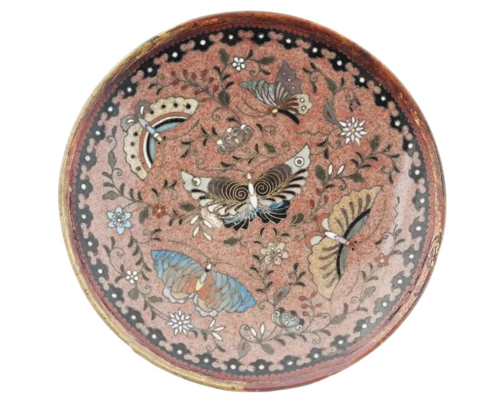 An antique Japanese Meiji period cloisonne enamel metal charger plate depicting butterfly images within floral designs. Features geometrical patterns to the border. The backside is adorned with a scrolling motif on a black ground with a central blue