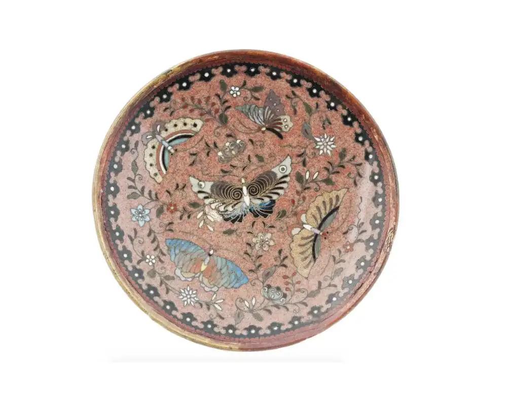 Antiguo plato de metal esmaltado en cloisonné japonés del periodo Meiji, con imágenes de mariposas dentro de diseños florales. Presenta motivos geométricos en el borde. El reverso está adornado con un motivo de roleos sobre fondo negro con un