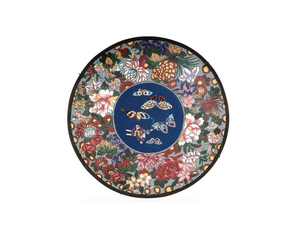 An antique Japanese Meiji period enamel charger plate. The plate is adorned with a central medallion featuring an image of butterflies surrounded by images of blossoming flowers made in the Cloisonne technique. The backside is adorned with a scaly