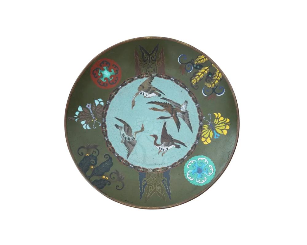 An antique Japanese late Meiji era decorative enamel over copper plate or charger. The interior of the plate is adorned with a polychrome design of flying geese surrounded by floral rondels and images of butterflies on the olive green ground on the