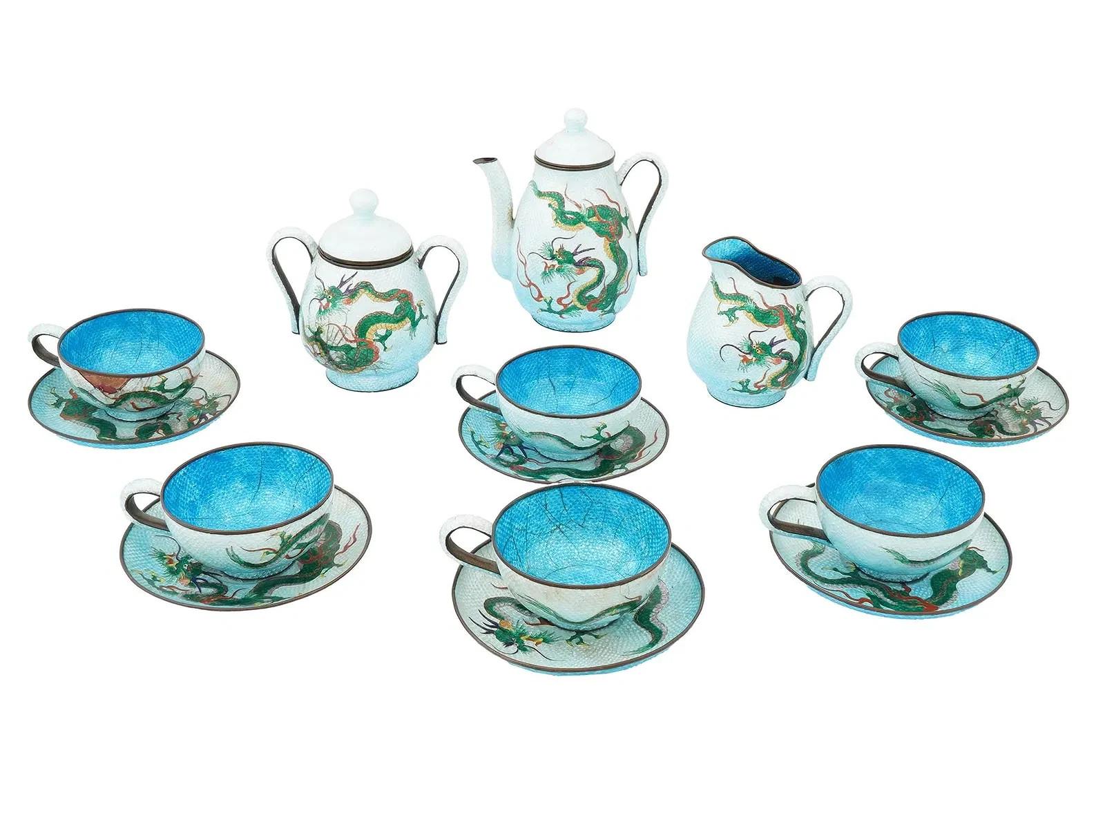 An antique Japanese Meiji period enamel tea set adorned with intricate dragon motifs. Consisting of six cups, six saucers, a creamer, a lidded sugar bowl, and a tea pot. The exterior of each piece has a white background adorned with green dragons.