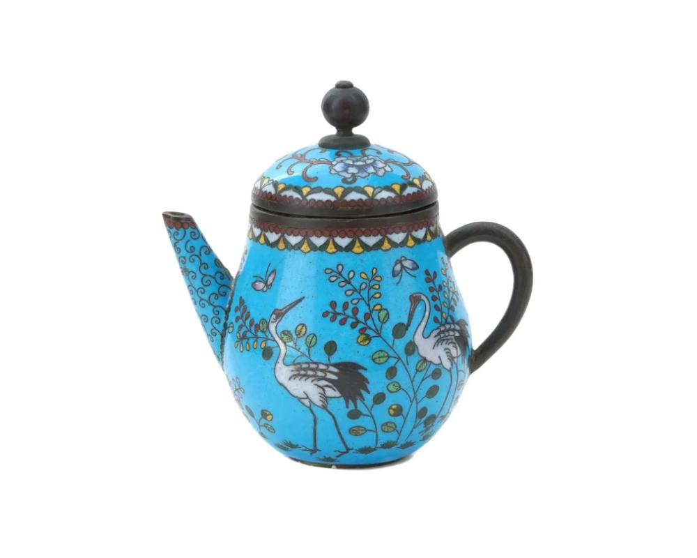 An antique Japanese, late Meiji era, lidded enamel over copper tea pot with a curved handle. The exterior of the ware is enameled with a polychrome design depicting cranes surrounded by plants and butterflies made in the Cloisonne technique. The lid