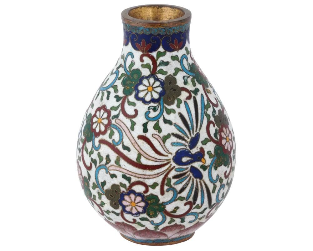 The globular shaped vase is adorned with polychrome enamel floral, foliage, and swirl motifs, and an image of a Phoenix bird on the white ground made in the Cloisonne technique. The base features swirl and lotus flower patterns made in the same