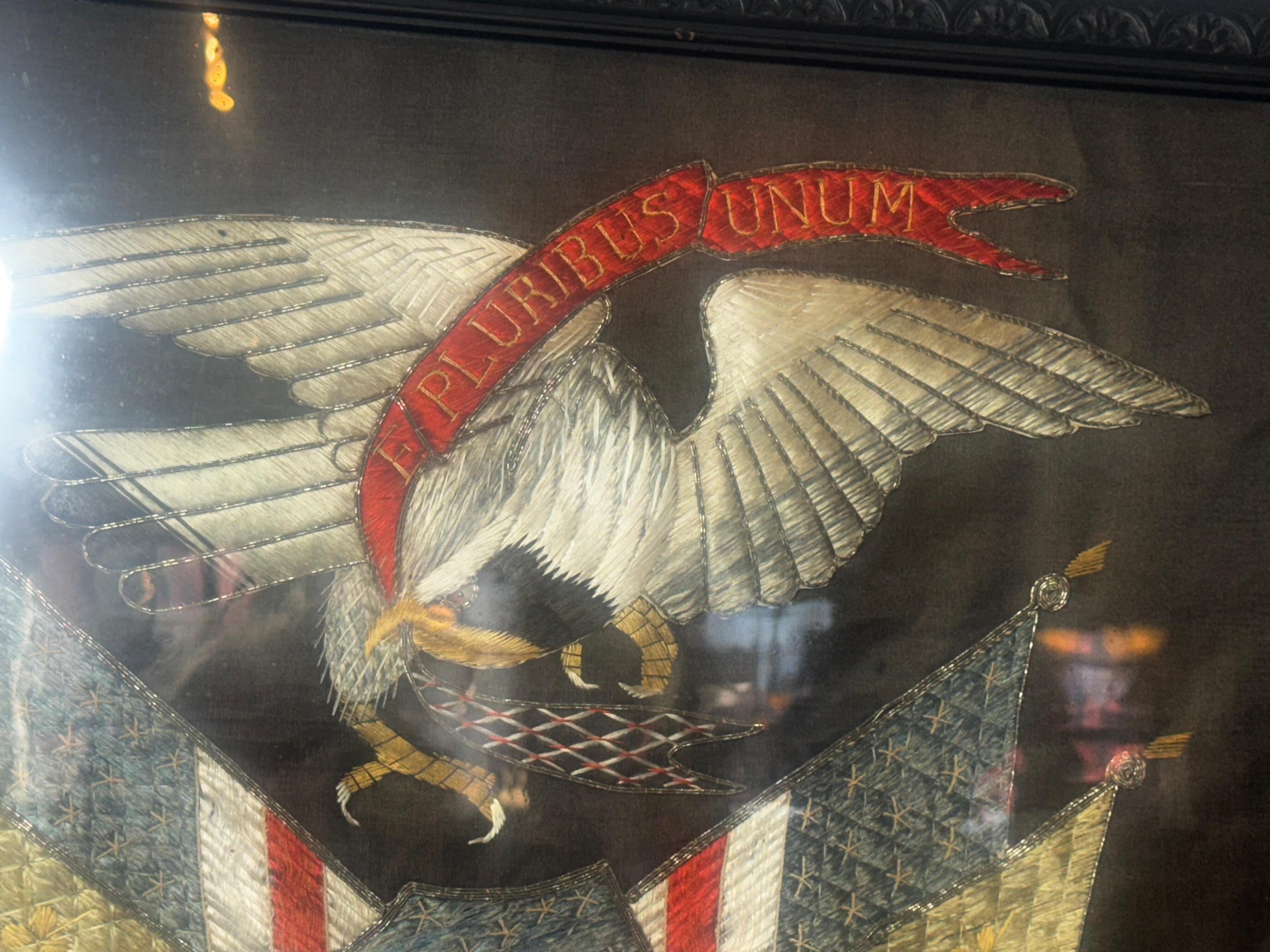 Un panneau de broderie en soie qui représente un aigle américain perché sur le bouclier de la liberté, avec des branches de laurier et des flèches de tir sous les pieds et flanqué de quatre drapeaux et bannières américains au dos. et la devise 