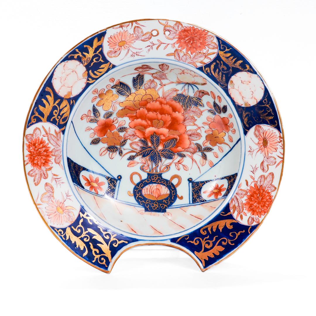 A fine antique barber's bowl.

In Japanese Imari porcelain.

With traditional blue underglaze and cold-painted iron red decoration and embellished with gilt highlights througout. The bowl's center depicting a large vase and peony flower in a field