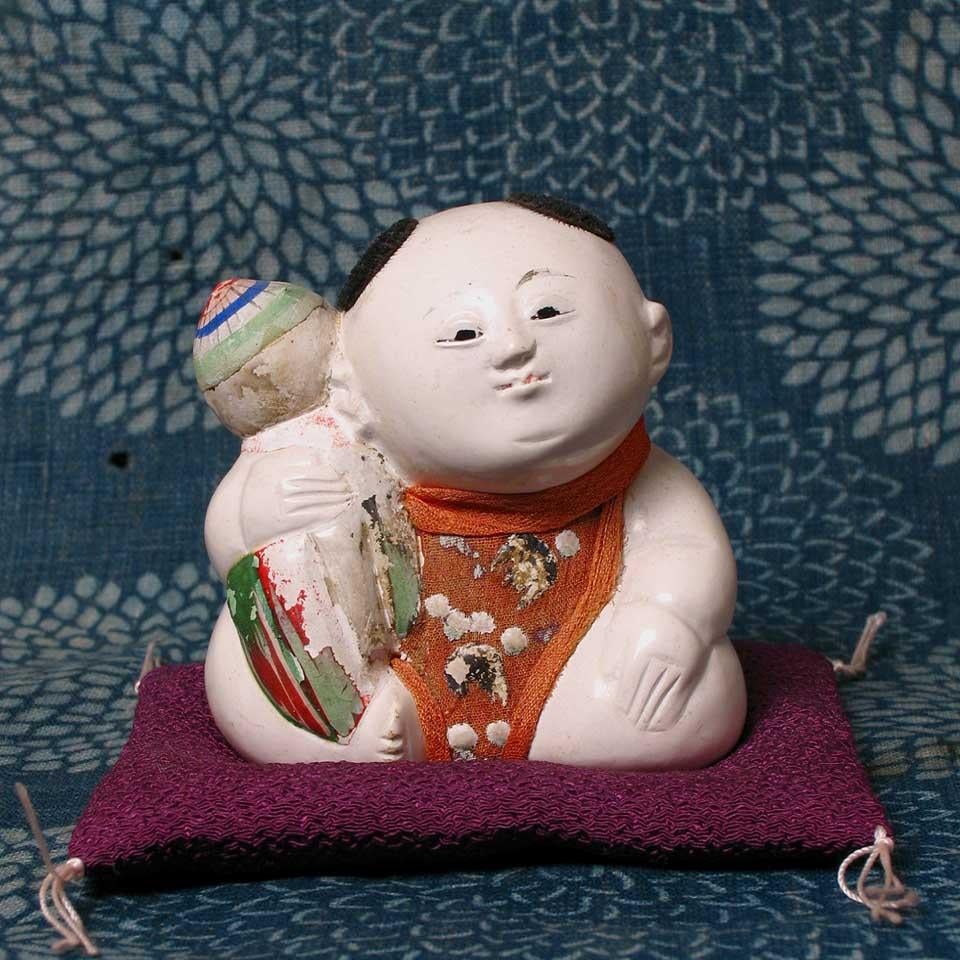 Japanese Kofuku-no-inori gosho ningyo (a good luck wish palace doll) of a plump male child seated with one leg tucked under and the other bent forward, holding symbols of mythical treasures the chintamani and the treasure cloak, traditionally given