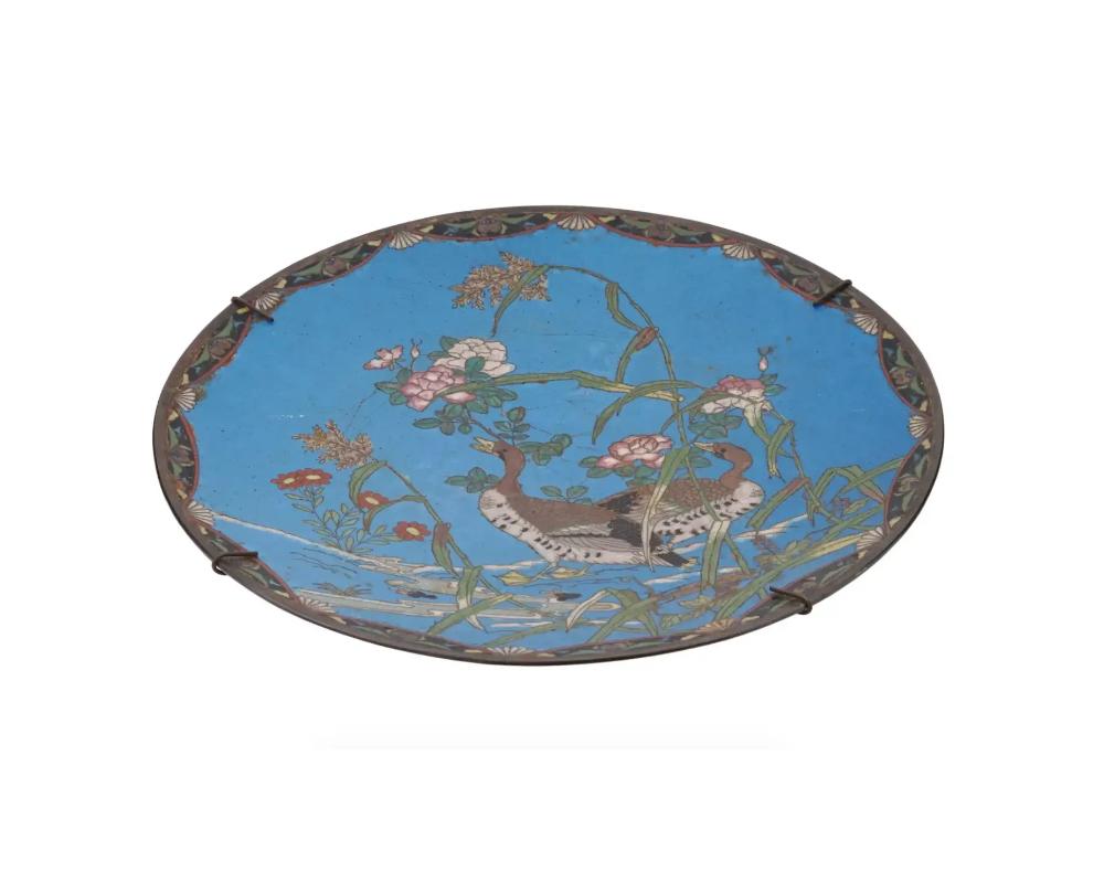 An antique Japanese late Meiji era enamel over metal charger plate. The plate is enameled with a polychrome image of naturalistic ducks sitting on branches with blossoming flowers on blue ground made in the Cloisonne technique. The border is