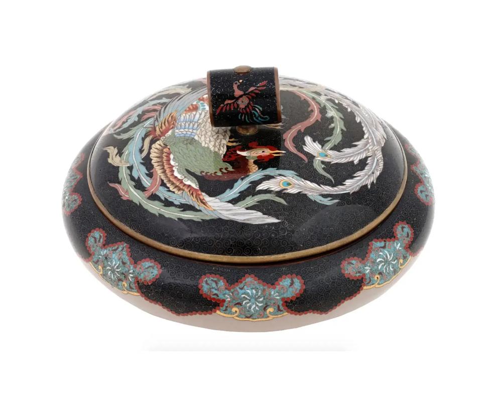 An antique Japanese Meiji Era covered enamel over brass jar. The cover of the jar is adorned with a polychrome image of a Phoenix bird surrounded by a swirl motif made in the Cloisonne technique. The cover is adorned with a tubby shaped handle,