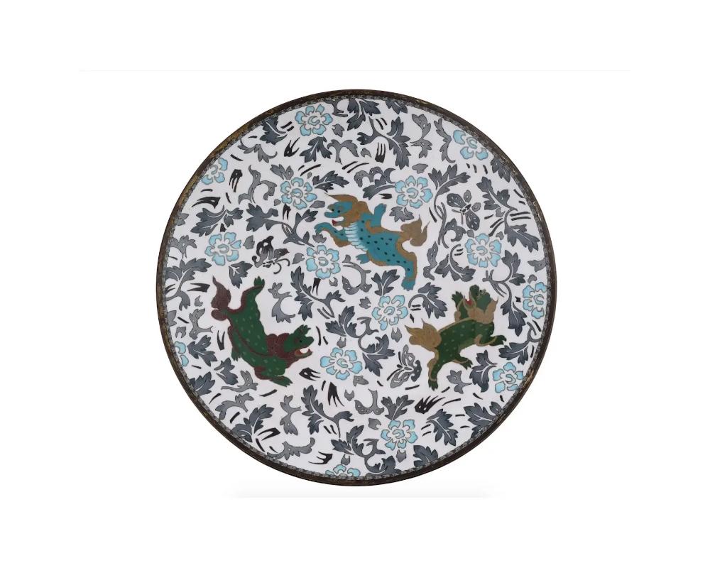 An antique Japanese Meiji era enamel over brass charger plate. The plate is enameled with a polychrome image of Foo dogs surrounded by a blossoming flower pattern on white ground made in the Cloisonne technique. The backside is adorned with a swirl