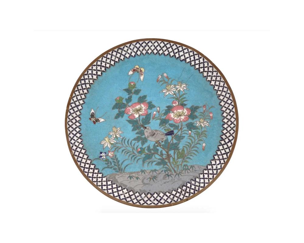 An antique Japanese Meiji era enamel over brass charger plate. The plate is enameled with a polychrome image of naturalistic butterflies and a bird sitting on a branch in blossoming flowers on blue ground made in the Cloisonne technique. The border