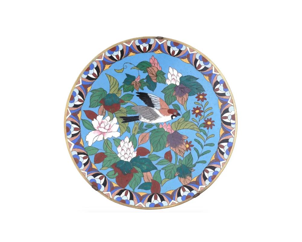 A highly decorative antique Japanese Meiji era brass plate. The exterior of the plate is adorned with a polychrome image of a bird flying among blossoming flowers and plants on a turquoise ground made in the Cloisonne technique. The border is