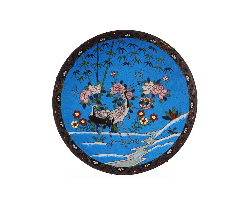 A high quality antique Japanese Meiji era decorative brass plate. The exterior of the plate is adorned with a polychrome image depicting two cranes in a landscape with blossoming flowers, bamboo trees, and plants on a turquoise ground made in the