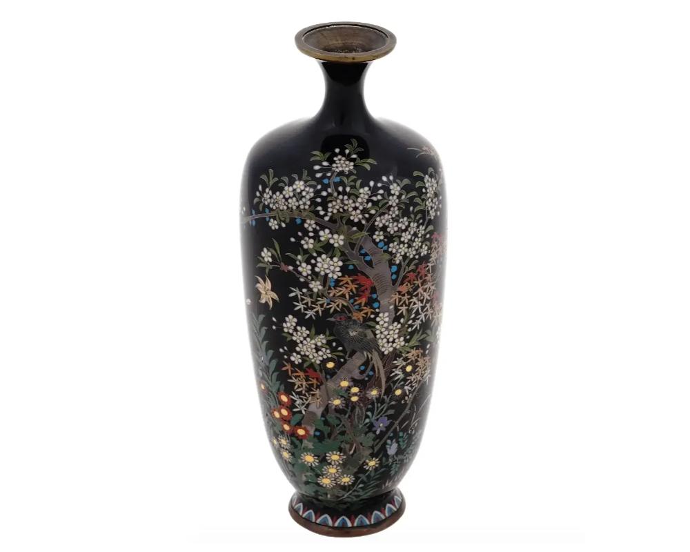 An antique Japanese, late Meiji period, enamel over brass vase. The vase has an amphora shaped body and a narrow fluted neck. The ware is enameled with a polychrome image of a bird in blossoming flowers and trees on the black ground made in the