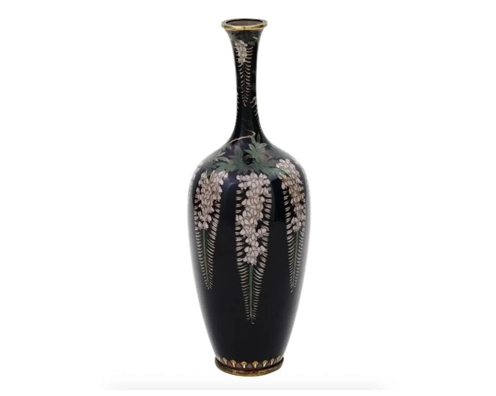 An antique Japanese, late Meiji period, enamel over brass vase. The vase has an amphora shaped body and a tall narrow neck. The ware is enameled with a polychrome image of wisteria blossoming flowers on the black ground made in the Cloisonne