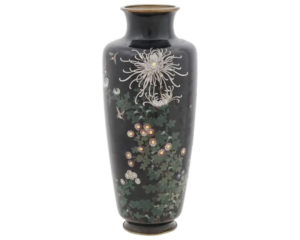 An antique Japanese, late Meiji period, enamel over brass vase. The vase has an amphora shaped body and a fluted neck. The ware is enameled with a polychrome image of birds in blossoming chrysanthemums and other flowers on the black ground made in