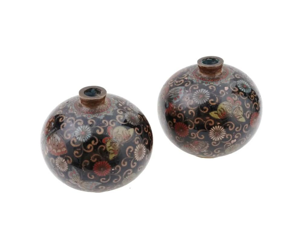 A pair of identical antique Japanese late Meiji era sphere shaped enamel over brass vases with narrow necks. The exterior of the vases are enameled with polychrome images of flowers and butterflies, surrounded by foliage and scrollwork patterns,