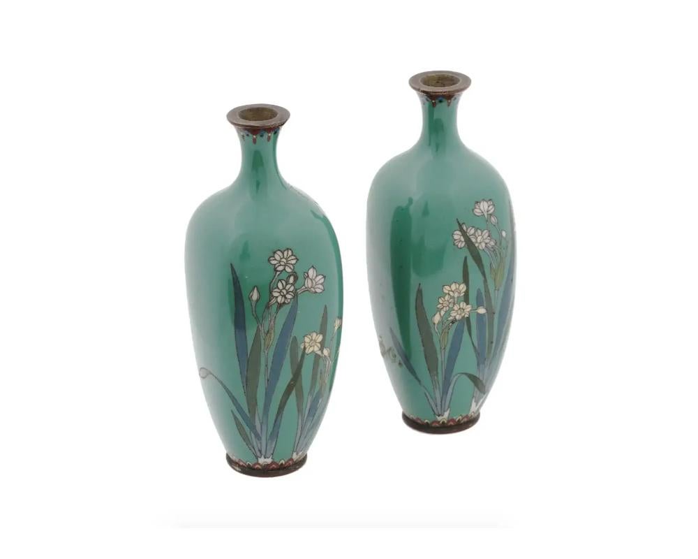 A pair of symmetrical antique Japanese Meiji period enamel over brass vases. Each vase has an amphora shaped body and a narrow neck. Each vase is enameled with a polychrome image of blossoming flowers on jade green ground made in the Cloisonne