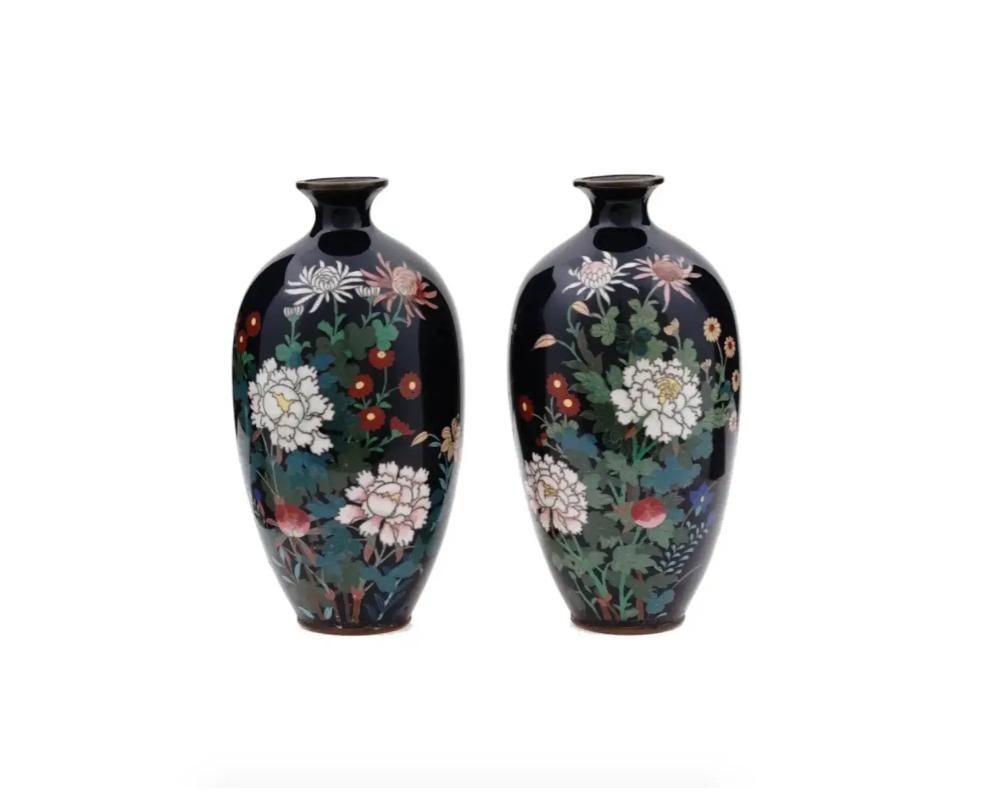 A pair of symmetrical antique Japanese Meiji period lobed enamel over brass vases. Each vase has an amphora shaped body and a narrow neck. Each vase is enameled with a polychrome image of butterflies in blossoming flowers surrounded by foliage,