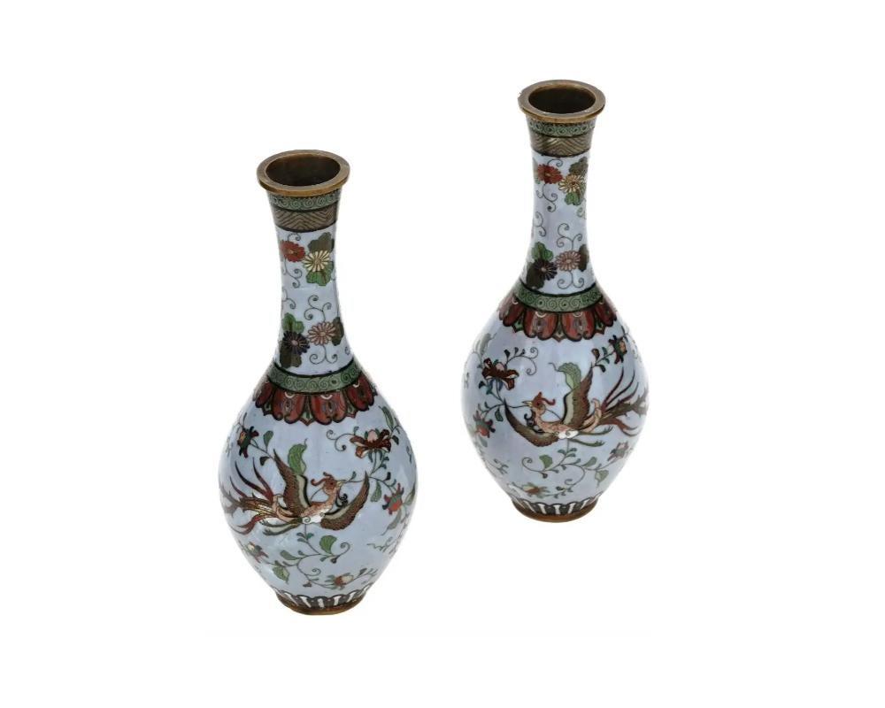 A pair of symmetrical antique Japanese, late Meiji period, enamel over brass vases. Each vase has a globular shaped body and a narrow fluted neck. Each ware is enameled with polychrome images of Phoenix birds surrounded by floral and foliage motifs