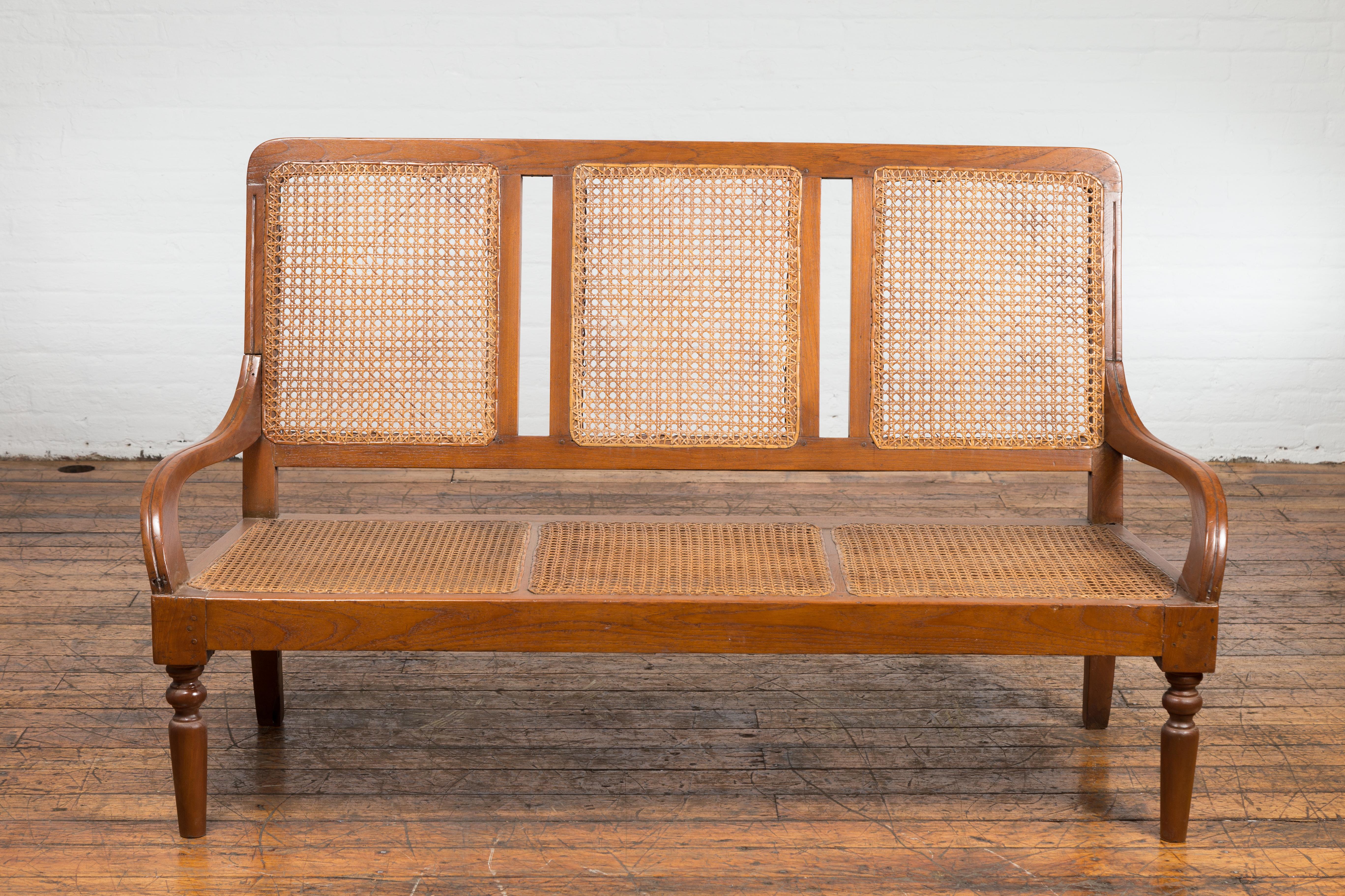 A rustic Javanese teak wood three-seat settee from the Turn of the Century with inset woven rattan seat and back. Infusing an air of rustic charm and understated elegance, this Javanese teak wood three-seat settee, hailing from the Turn of the