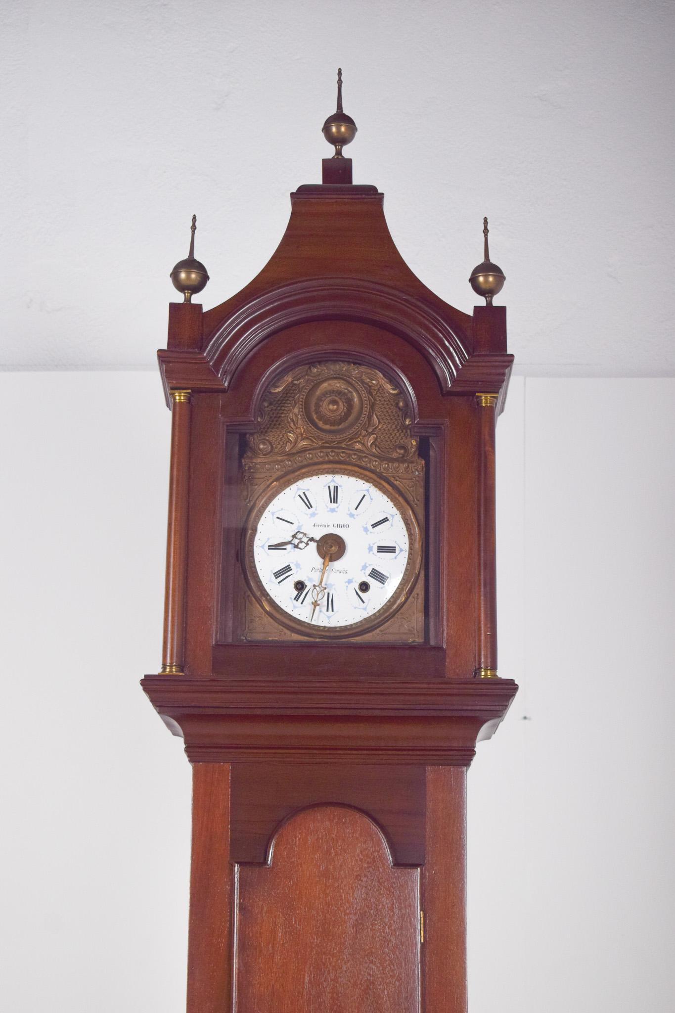 Großvateruhr von Jérémie Girod, Porto und Coruña. Die Struktur ist aus dunklem Holz, mit einer polierten Oberfläche und eleganten Linien. Im oberen Teil befindet sich das runde Zifferblatt mit römischen Ziffern und verzierten Zeigern, eingefasst in