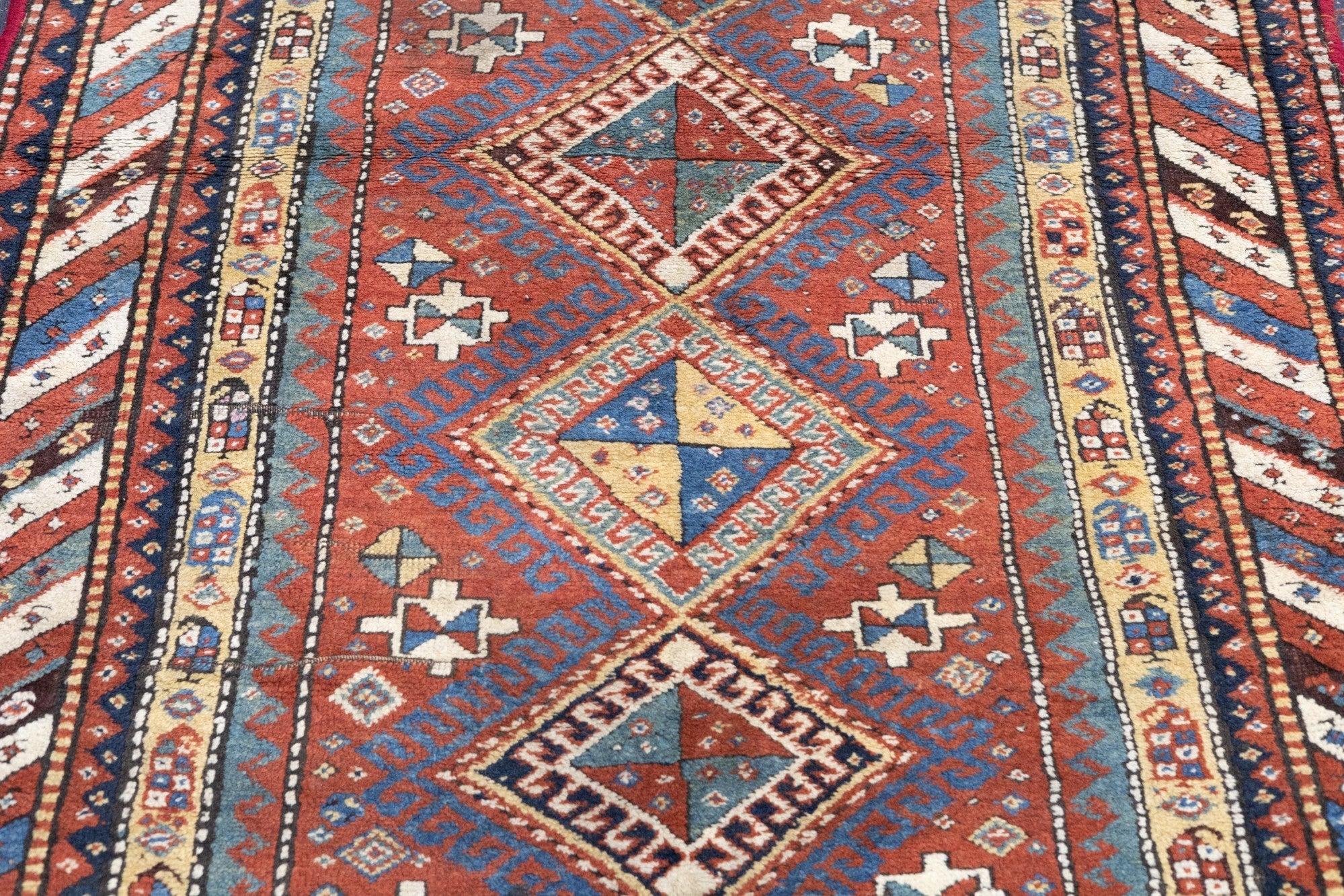 Tapis Kazak antique 3'10'' x 6'11''. Le champ rouge présente un médaillon composé de cinq diamants à crochets de taille égale, de couleur bleue, crème, verte et camel. Carrés secondaires avec des extensions courtes. Bordure principale rayée en