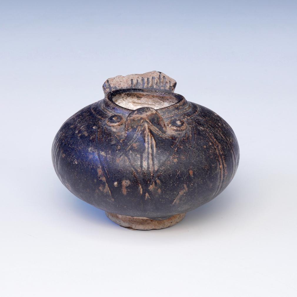 Khmer Ceramic Lime Pot. A squat globular stoneware jarlet in the form of a bird covered in a thick dark brown glaze stopping short of the elevated string cut foot displaying the beige biscuit, a large owl’s beak and protruding eyes in relief on the