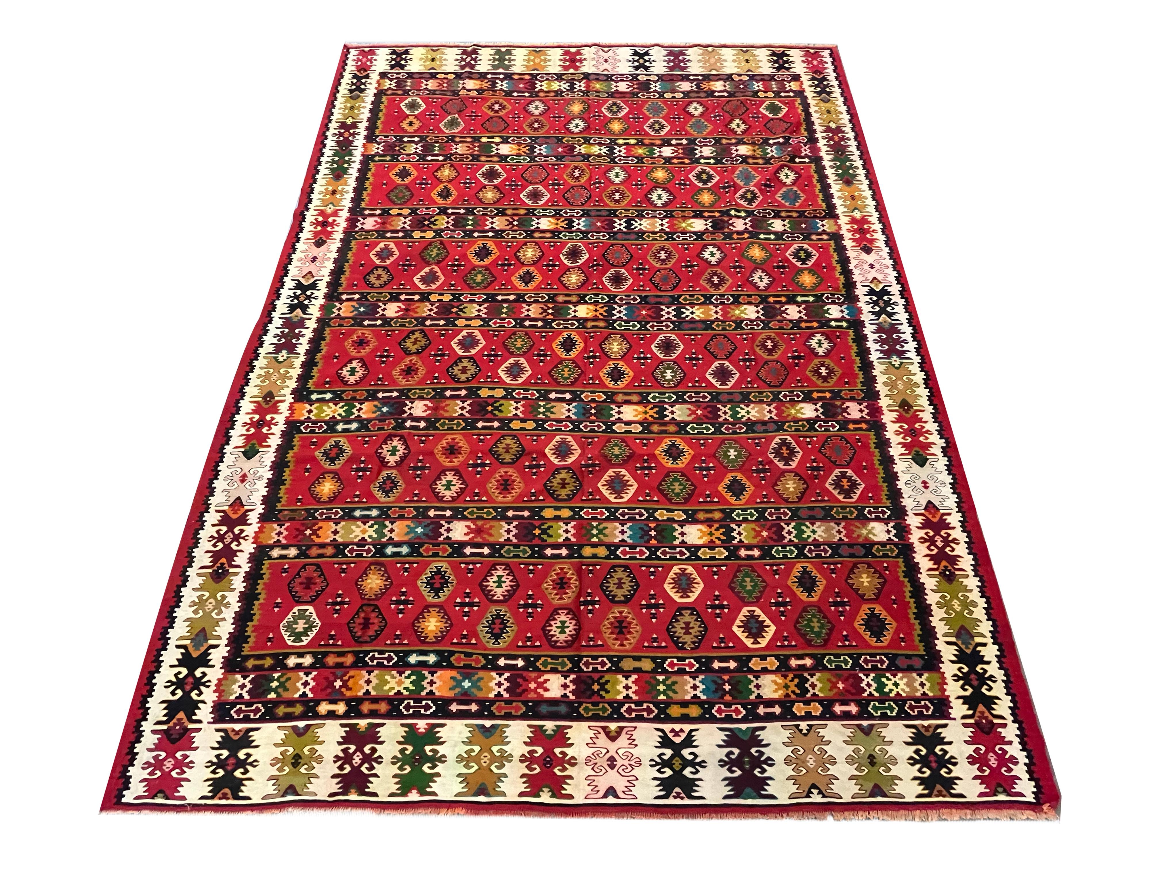 This bold red kilim is a traditional flatwoven area rug woven by hand circa 1910. The design features a geometric design woven in bold accents of black orange green and various other colours on a bold red background. The stripe design is eyecatching