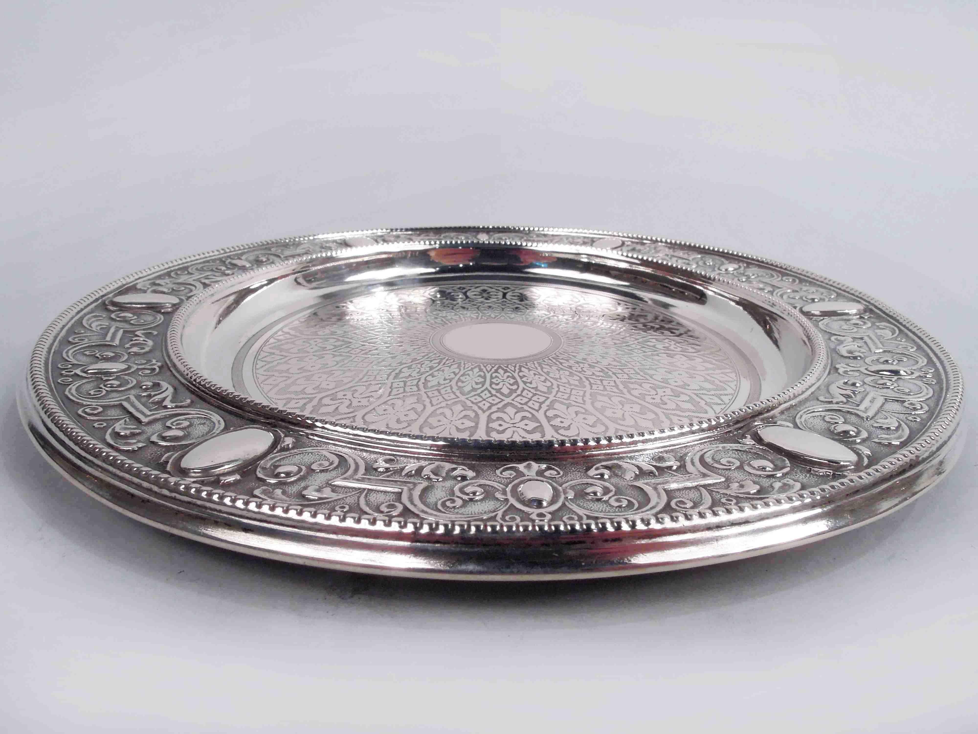 German Renaissance 800 silver wine bottle coaster, ca 1890. Round and deep well. Central rondel (vacant) in engine-turned frame radiating leaf and scroll ornament on lined ground. Wide and flat rim with embossed curvilinear scrollwork and ovals