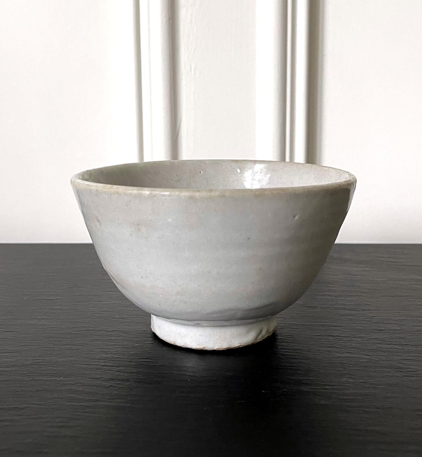 A Korean ceramic rice bowl covered in a white glaze with a hint of bluish green color circa 19th century toward the end of Joseon Dynasty. The deep bowl was thickly potted and made for everyday use, featuring a classic form with a short cylindrical