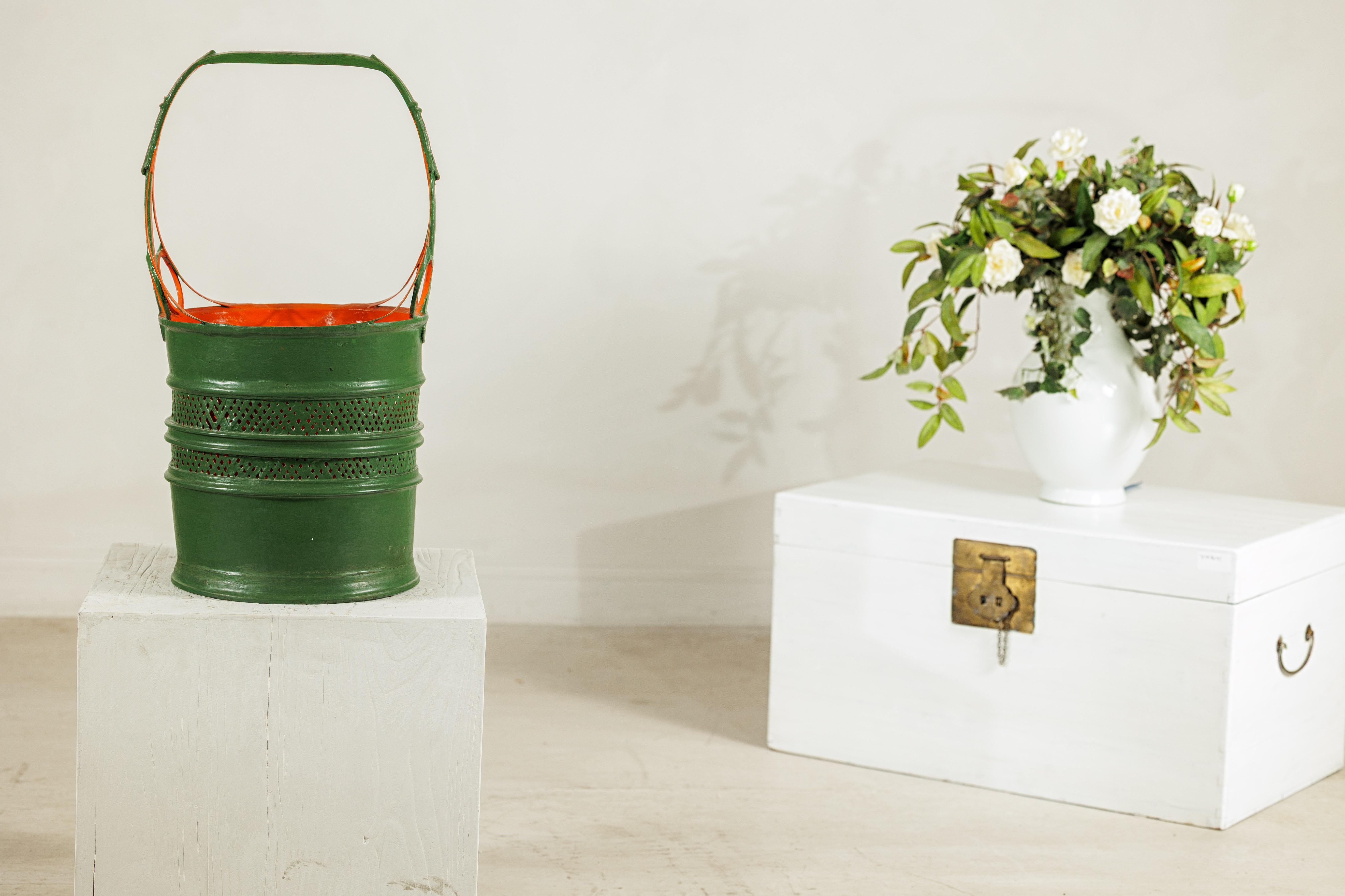 Ce remarquable panier en métal antique se distingue par sa finition laquée bicolore éclatante et sa silhouette sculpturale. L'extérieur vert profond contraste magnifiquement avec l'intérieur orange vif, créant un couple de couleurs audacieux mais