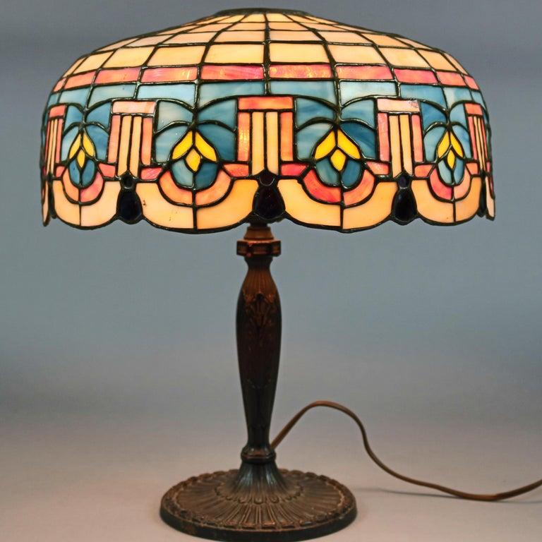 An antique Arts & Crafts table lamp by Lamb Brothers offers domed leaded slag glass shade with stylized foliate and tassel decoration having scalloped edge and surmounting cast single socket base with maker stamp as photographed, circa