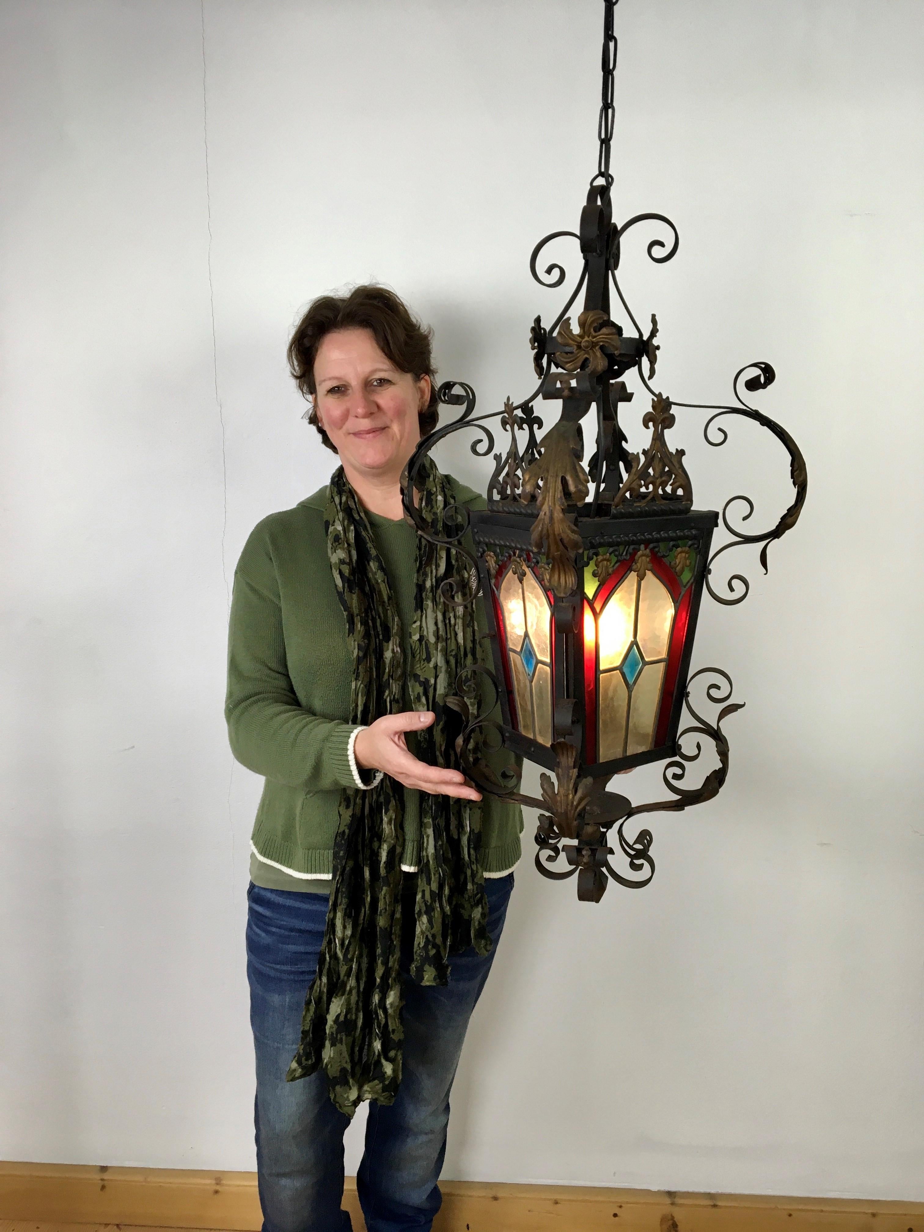 Antique lantern with colored stained glass - leaded glass 
A wrought iron French candle lantern from the Early 20th century.
The iron frame has a black painted finish with gold - bronze colored leaves, flowers  and details around. Stained glass in