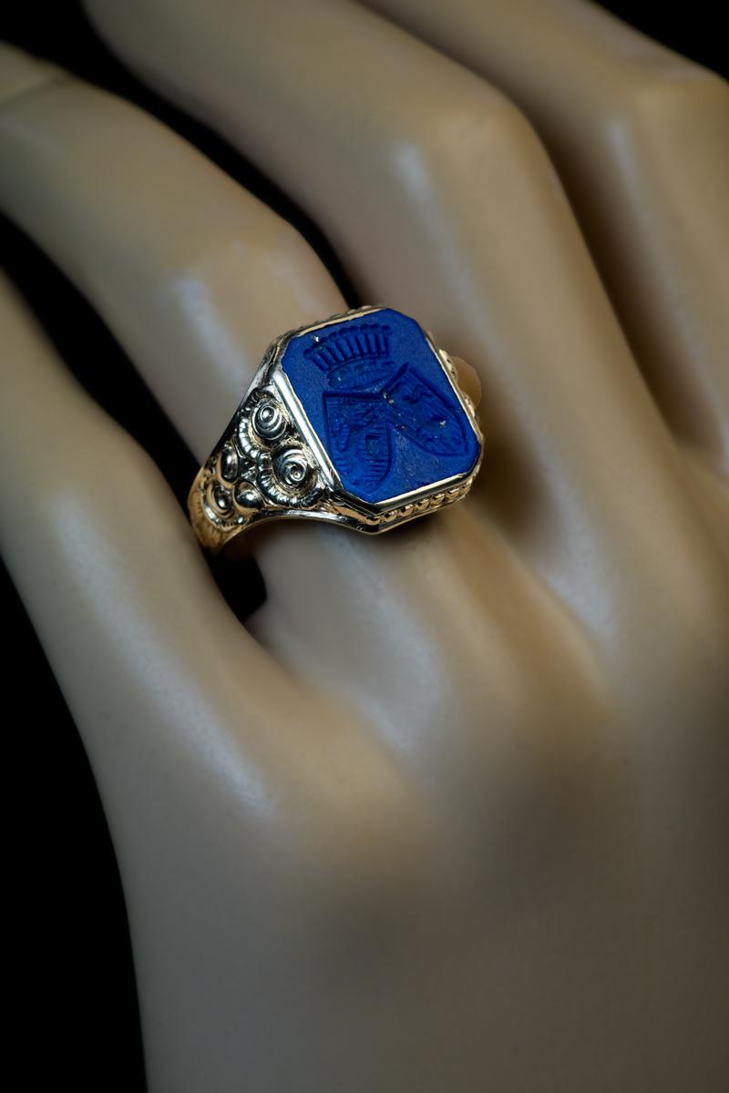 Circa 1900

The 14K gold unisex ring with stylized floral design on the shoulders features a lapis lazuli intaglio of an excellent purplish blue color with some fine golden inclusions of pyrite. The lapis matrix is engraved with a coat of arms
