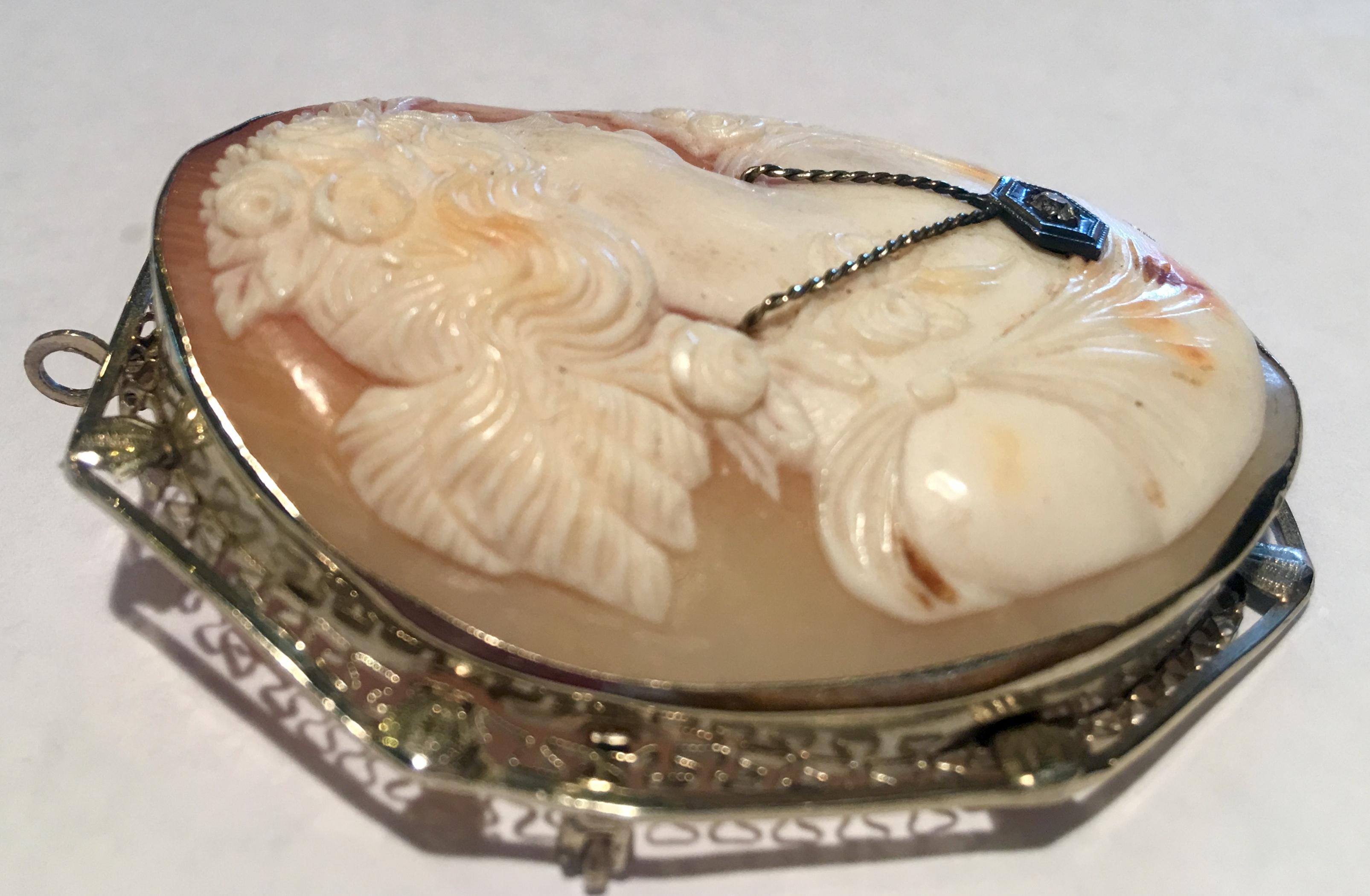 Antique, large, 14 karat white gold, elaborately hand carved shell cameo brooch or pendant depicts a woman in profile, with a pompadour and gown festooned with flowers and wearing a necklace with a single rose cut diamond.

May be worn as a brooch