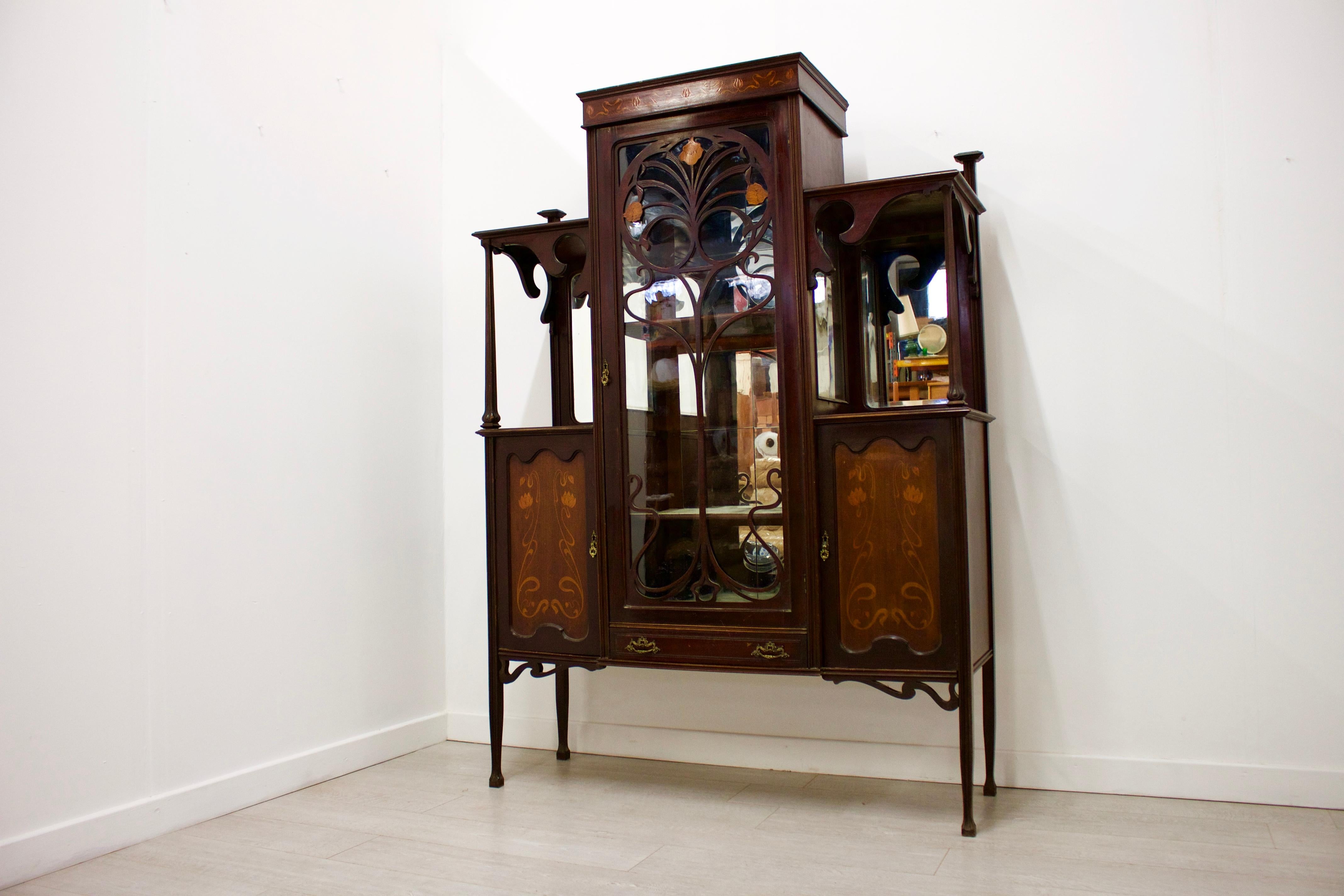 Art Nouveau display cabinet, in the manner of Shapland & Petter.
- Made from mahogany, inlaid with satinwood.
- Features central glazed cabinet, overlaid with a whiplash design
& Iris flower.
- Flanked by Marquetry panelled doors.