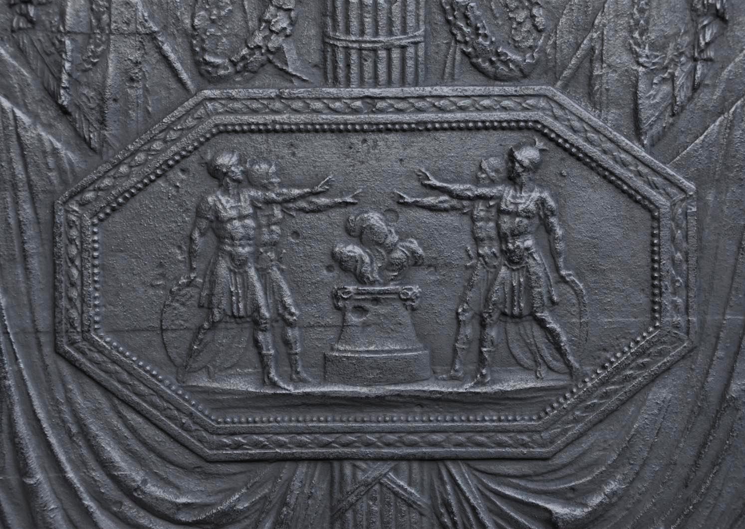 Cette plaque de cheminée ancienne a été fabriquée dans la première moitié du 19e siècle. Au centre, dans un médaillon octogonal, quatre soldats armés en tenue d'inspiration antique lèvent vigoureusement les bras au-dessus d'un feu. Leur pose est