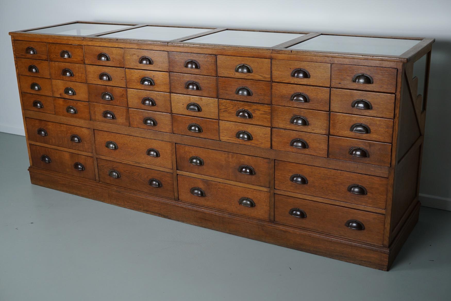 This oak art deco haberdashery shop counter dates from the 1920-1930s and was made in Amsterdam by Joh. Tacoma. It features a solid wooden frame, a glass casing with folding doors in the top and many drawers in oak with bronze handles.